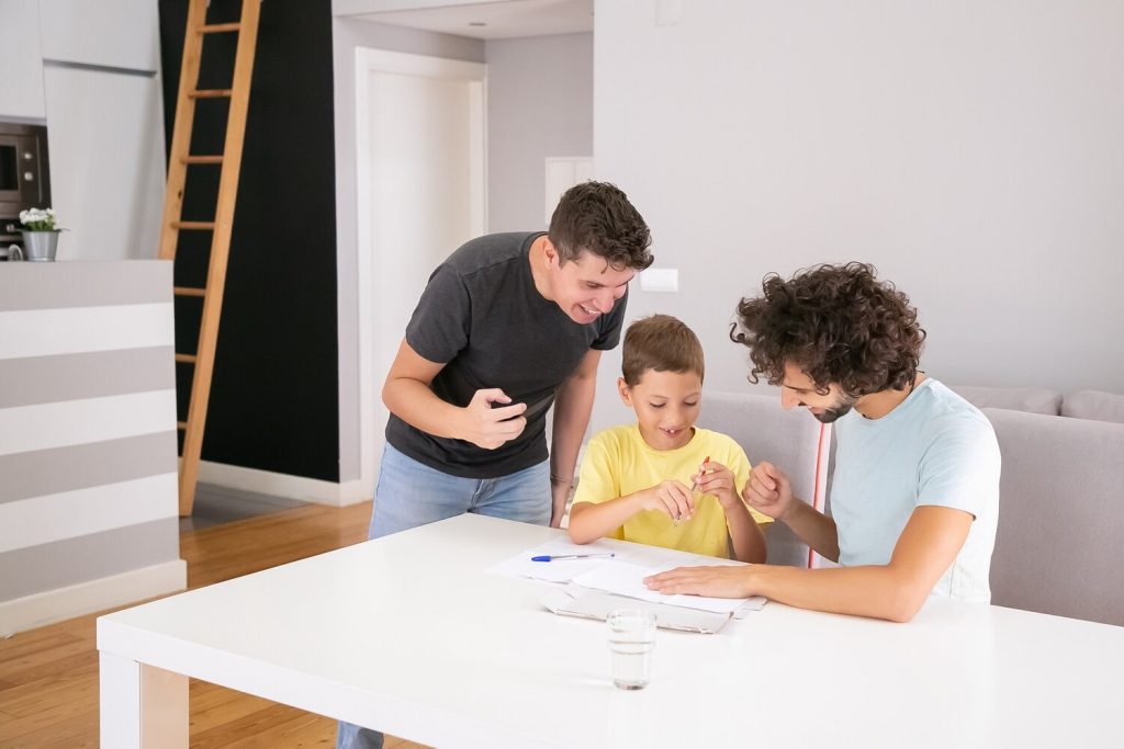 happy couple of dads helping focused boy with school home task, sitting at table together, writing on paper. family and gay parents concept