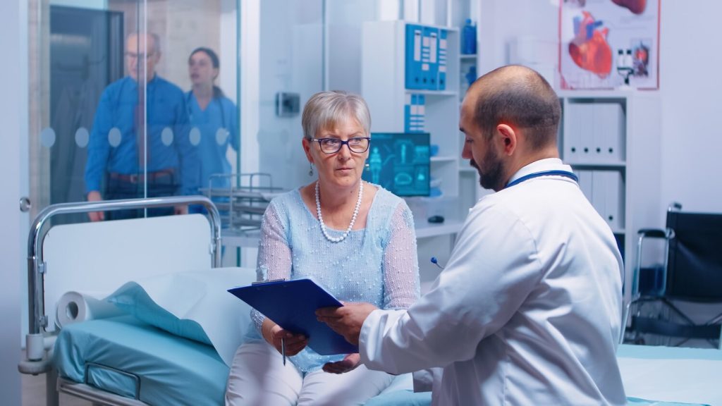 senior patient signing medical decisions form sitting on hospital bed in modern private clinic. doctor with clipboard, nurse working in backgorund. healthcare medical medicinal system documents contra