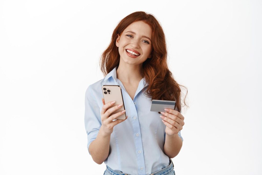happy smiling shopping girl holding mobile phone, credit card, paying in smartphone app, order something on site, checking bank account, standing against white background.