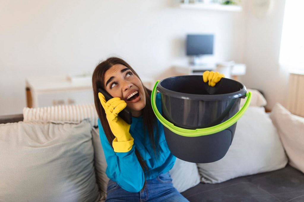 shocked woman calling plumber while collecting water leaking from ceiling using utensil