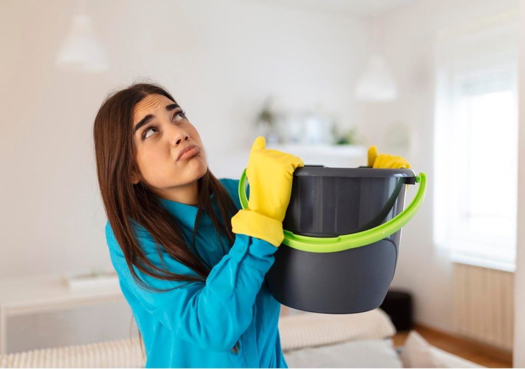 shocked woman looks at the ceiling while collecting water which leaks in the living room at home worried woman holding bucket while water droplets leak from ceiling in living room